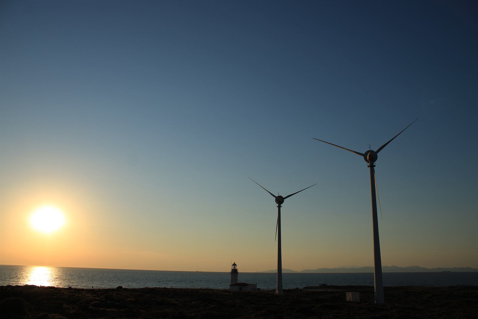 Two wind turbines and a lighthouse on the coast at sunset